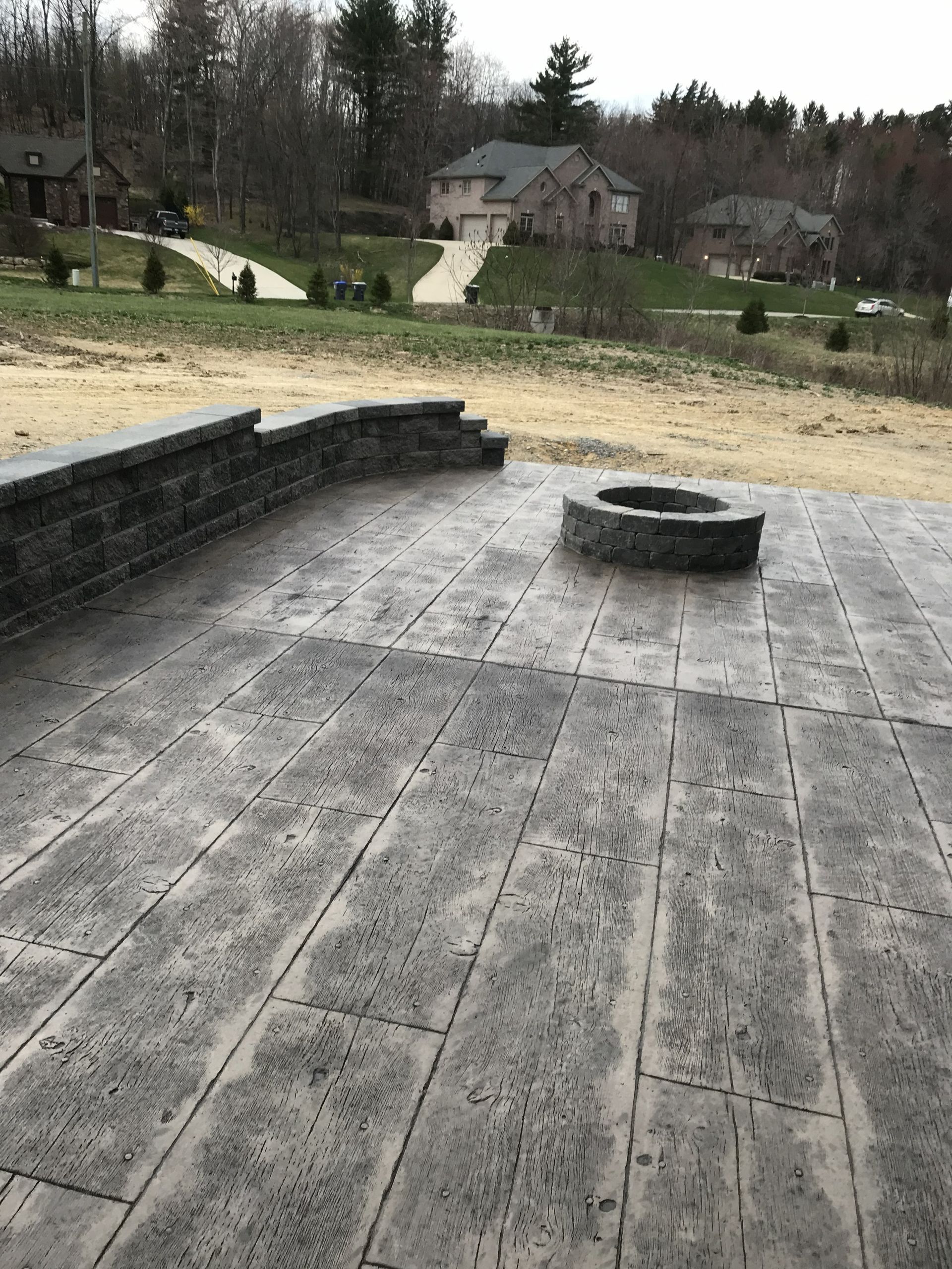 Stone patio with low curved wall and fire pit, overlooking a grassy yard and houses beyond.