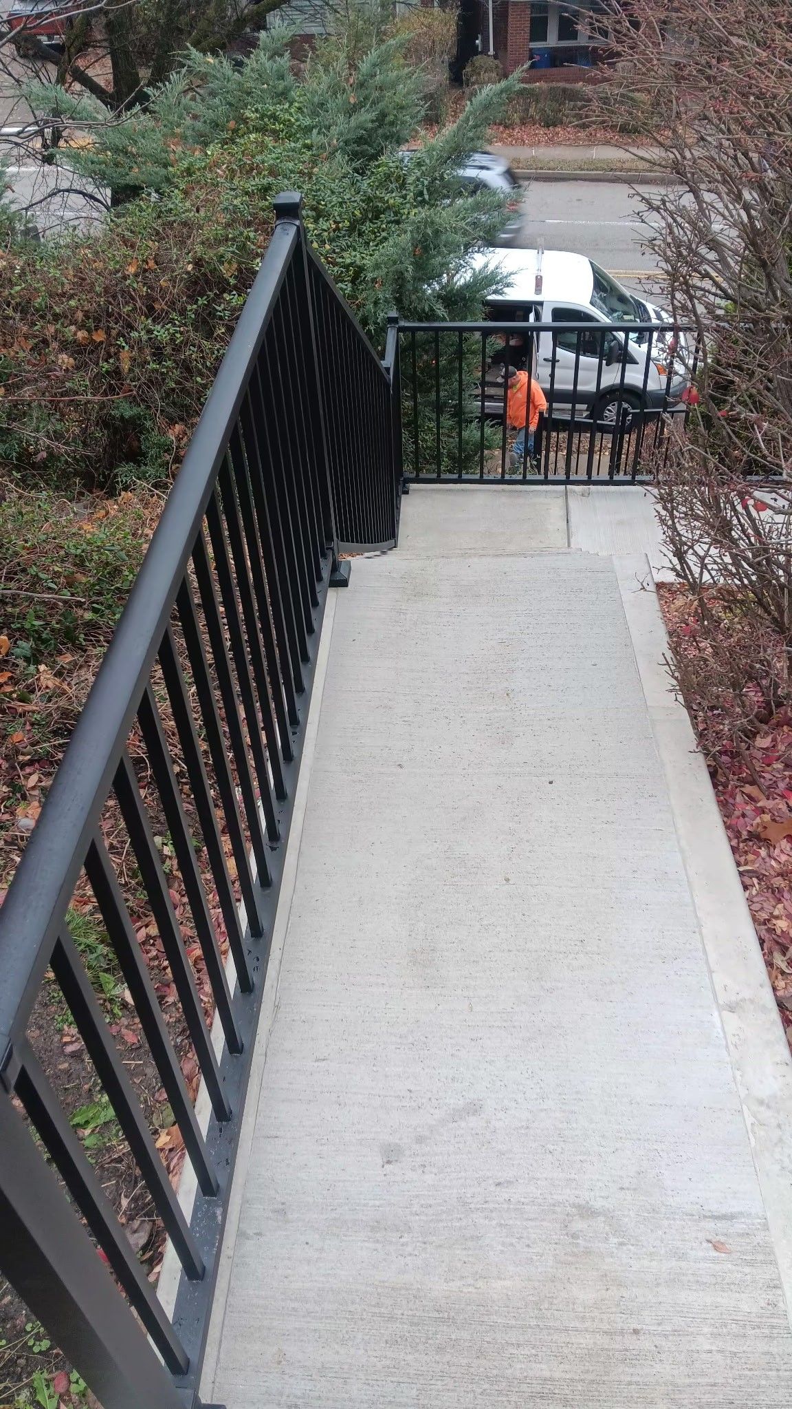 Narrow outdoor walkway with black railing, leading down to a gate and parked white van below.