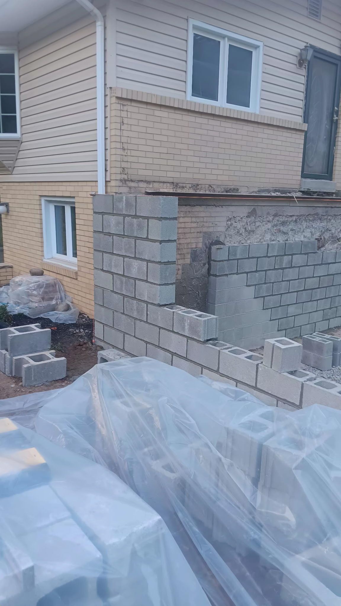 Partially built cinder block wall beside a beige house, with construction materials and plastic sheeting in the foreground