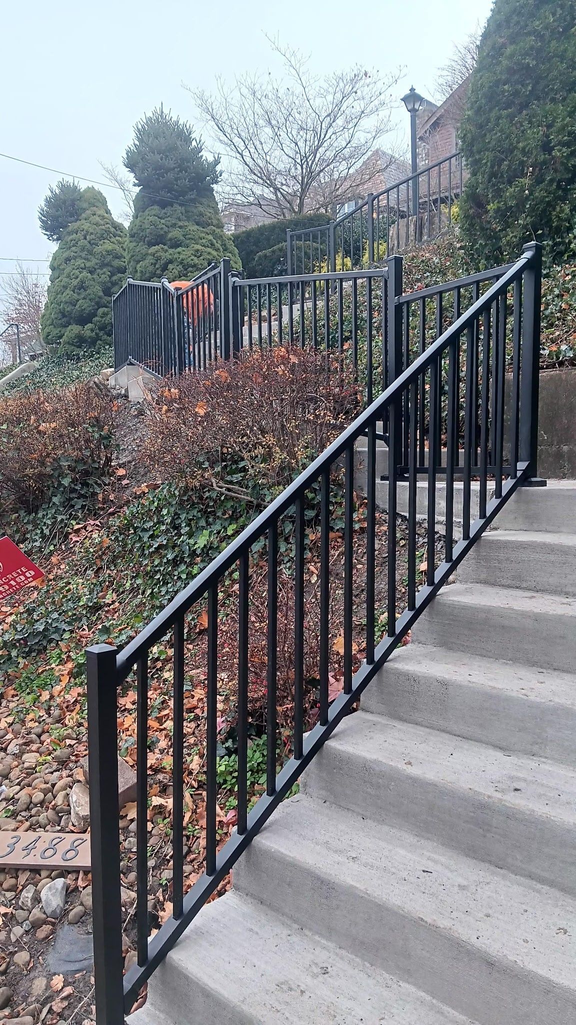 Concrete outdoor stairs with black metal railings beside shrubs and trees on a hillside
