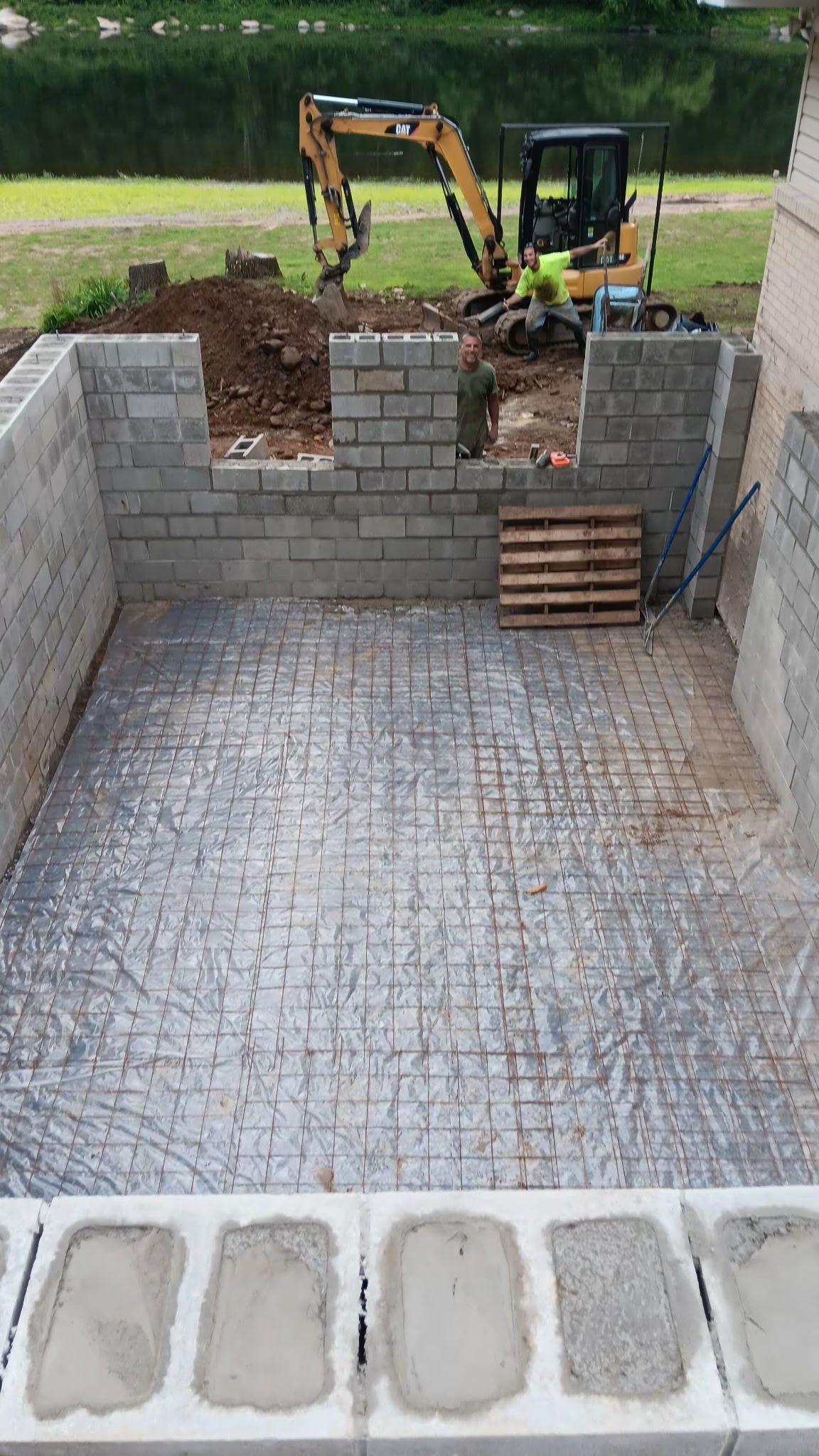 Excavator working behind a stone retaining wall above a paved lower area, with stacked bricks in front