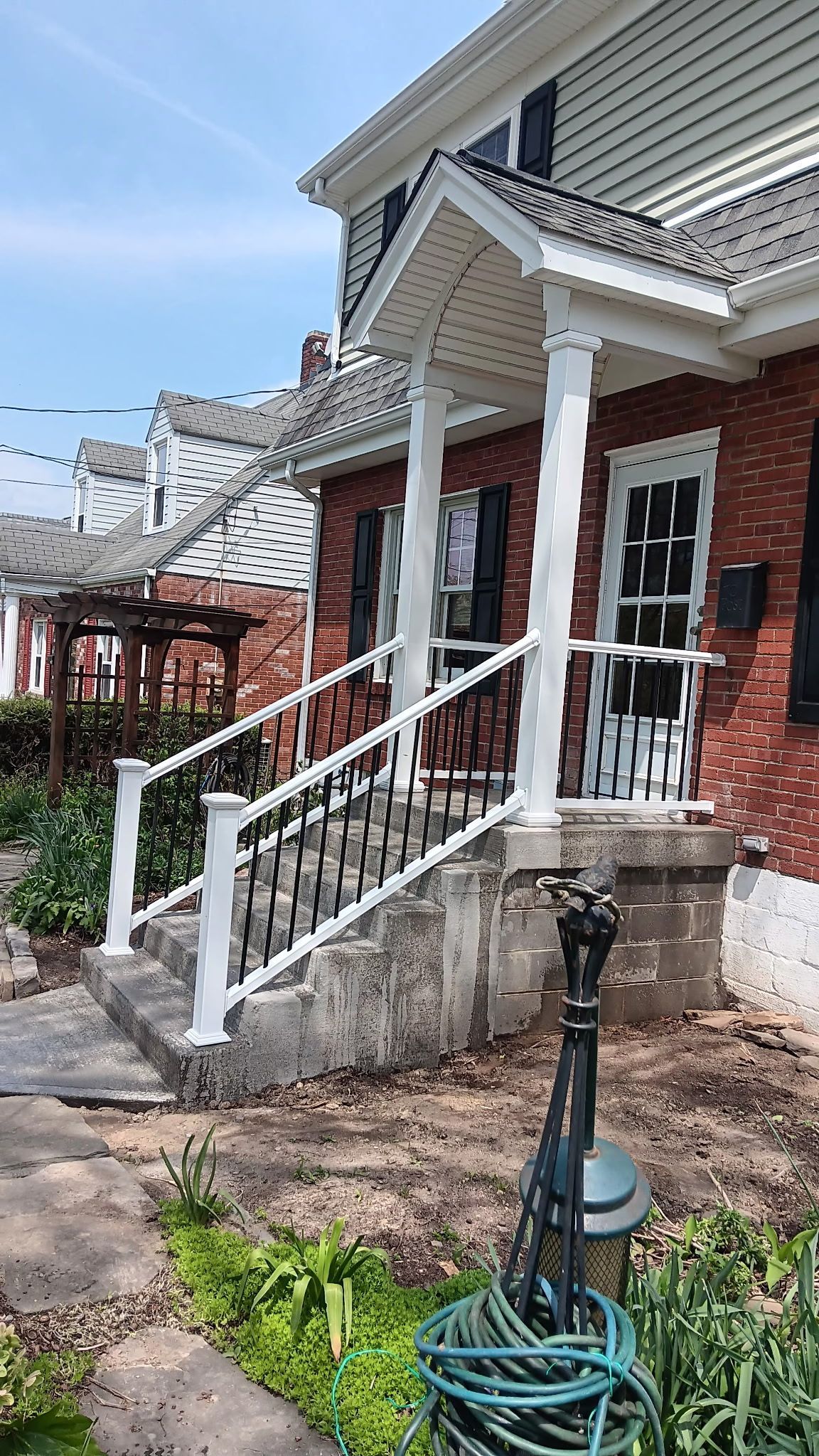 Front steps of a brick townhouse with white railings and a coiled green hose in the yard