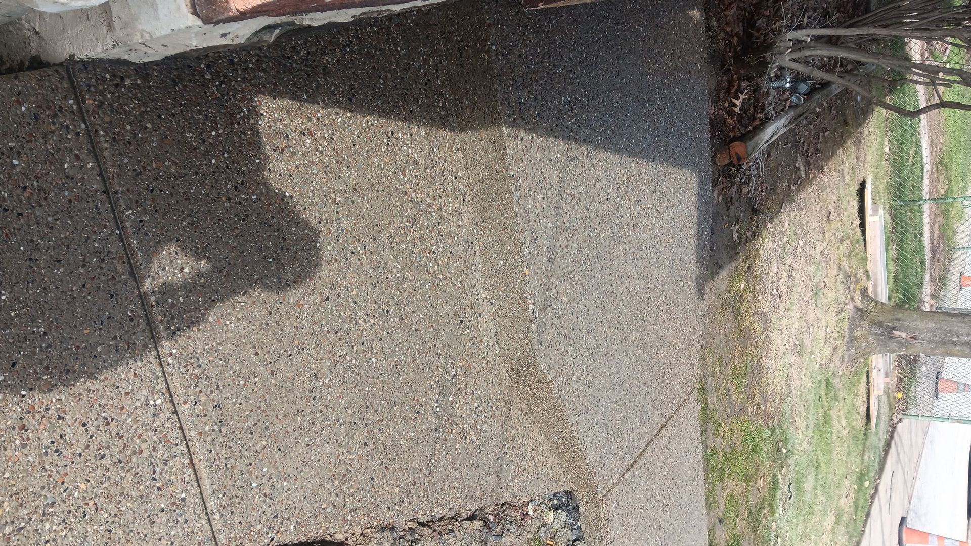 Wet concrete walkway beside a road, with a shadow cast across the surface and grass along the edge.