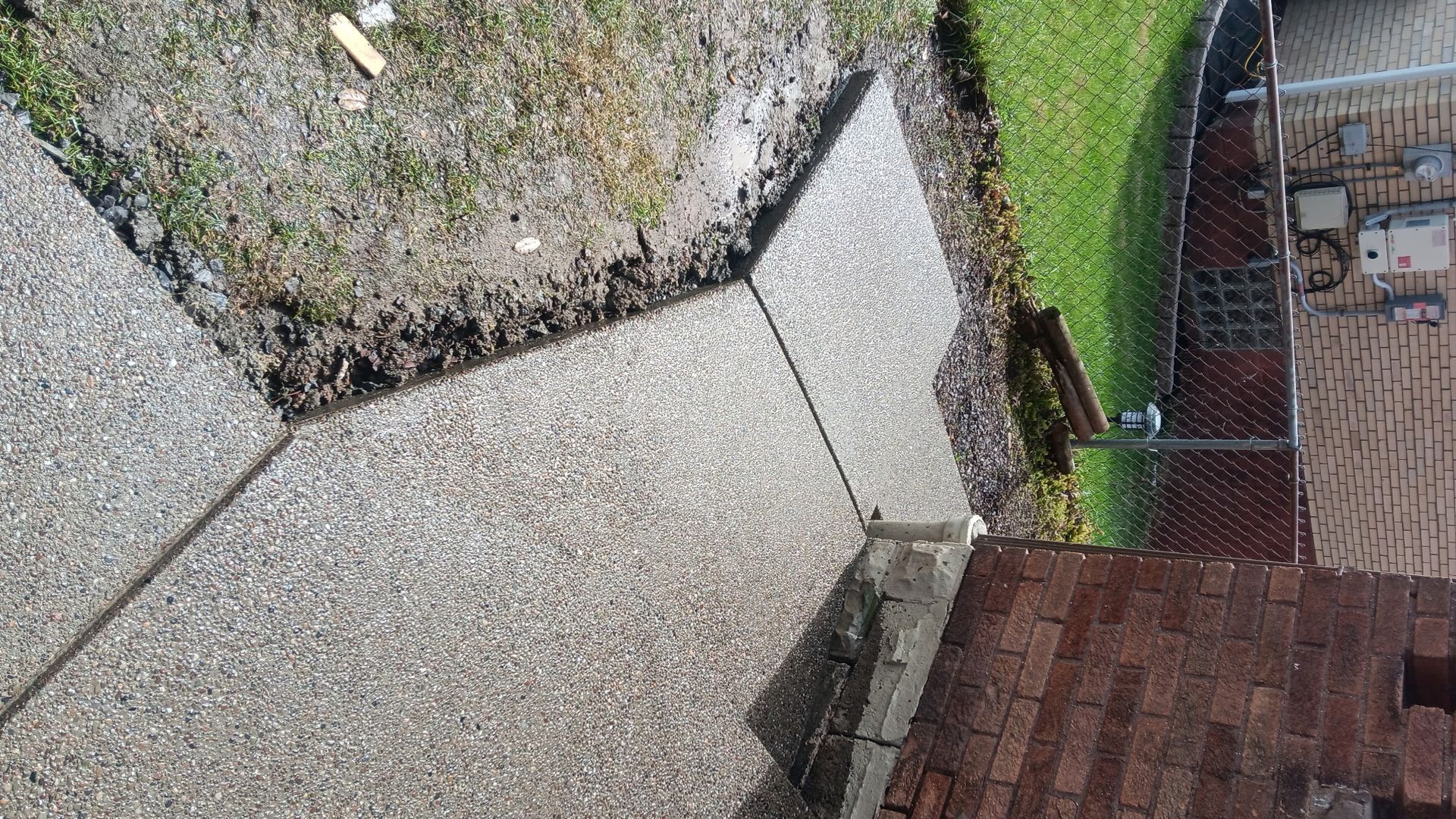 Concrete patio beside a gravel yard and red brick wall, with a gate and grass in the background.