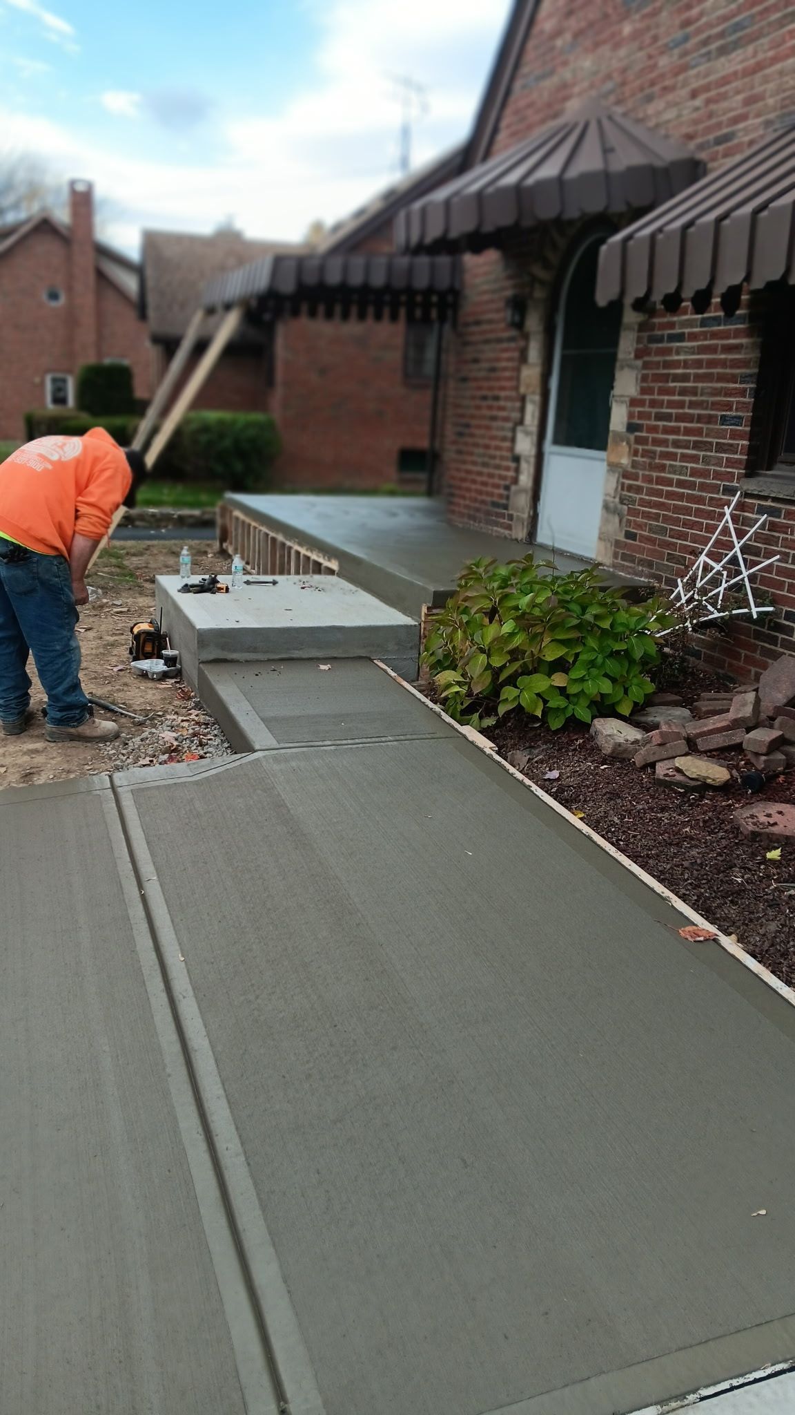 Worker near a brick building installs a concrete sidewalk ramp beside awnings and landscaping.