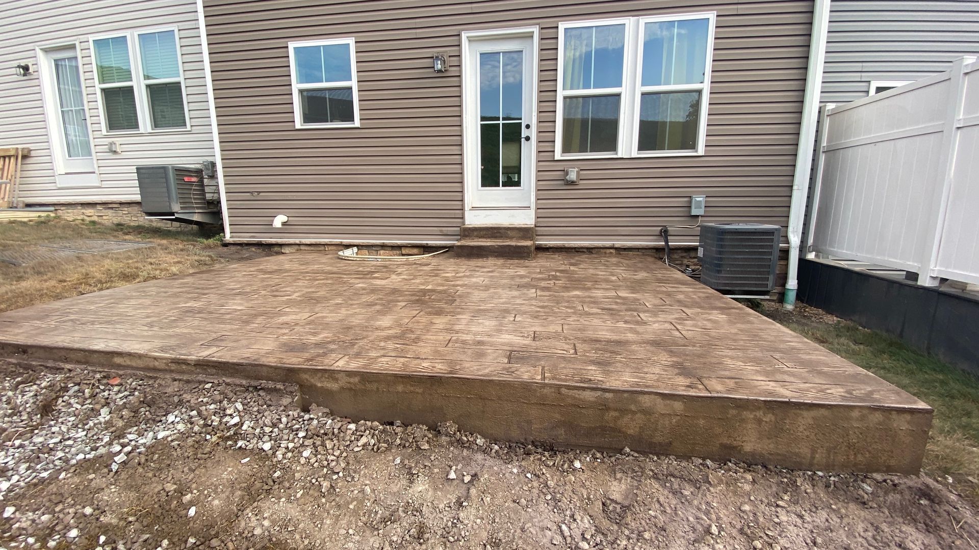 Concrete patio behind a brown house with a white door, windows, and a white fence on the right.
