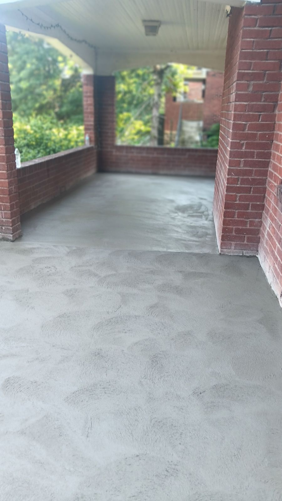 Covered brick porch with a smooth concrete floor and railing overlooking greenery