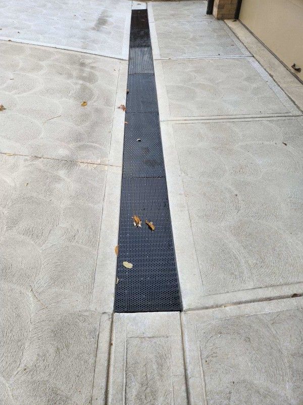 Sidewalk with a long narrow drainage channel and a few leaves on the pavement.