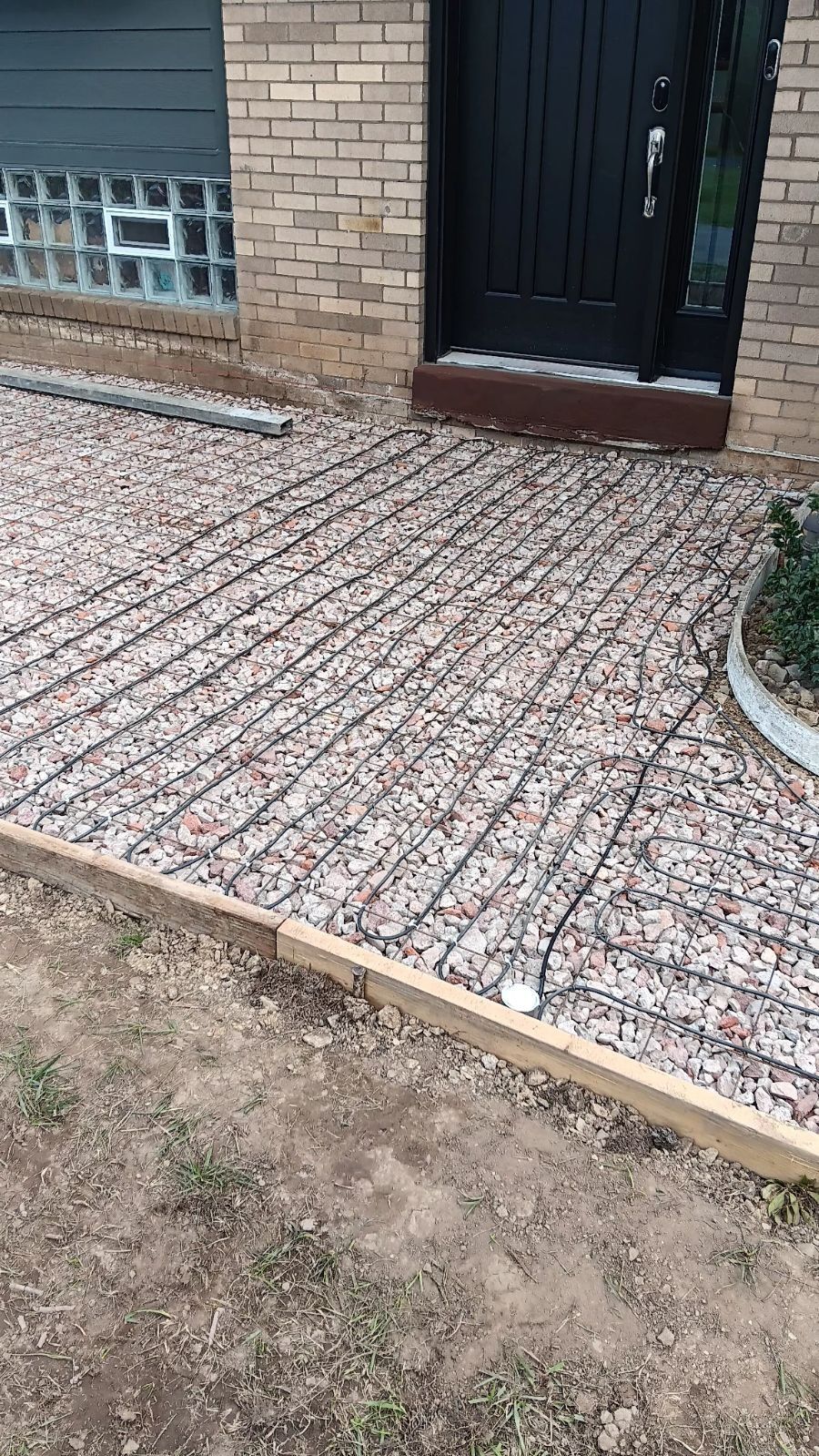 Brick house entrance with a black door and patterned tiled walkway beside a dirt yard.