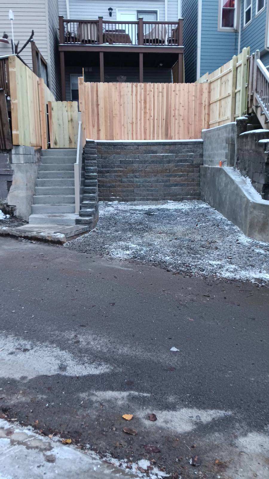 Steep retaining wall and stairs leading to a fenced backyard behind a house, with a snowy driveway below