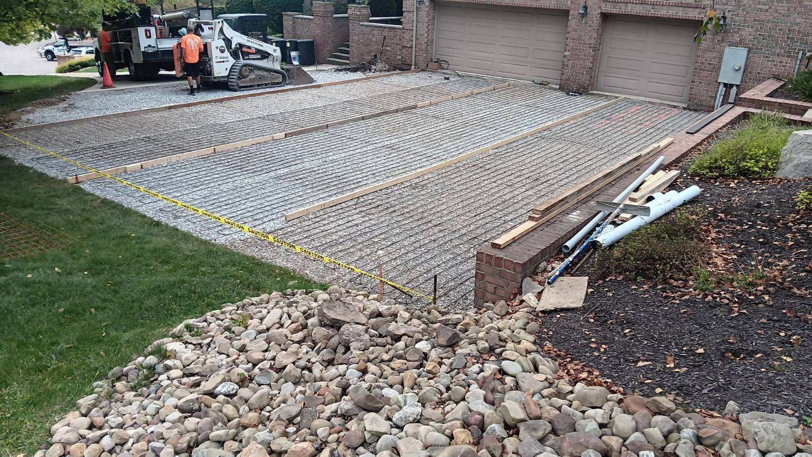 Driveway under construction with gravel base, rebar grid, and workers near a house.