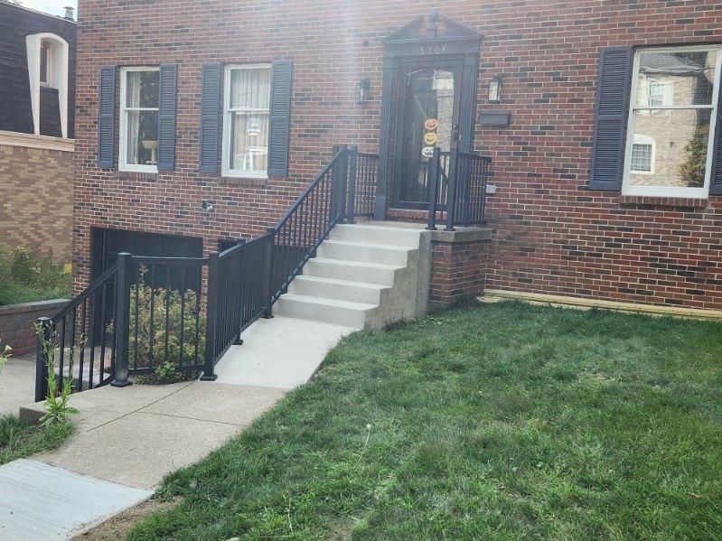 Brick apartment entrance with concrete steps, black railing, and a small green lawn.