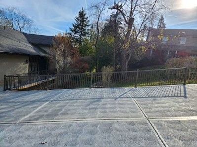 Concrete driveway beside a sloped metal fence, trees, and a dark building in bright daylight