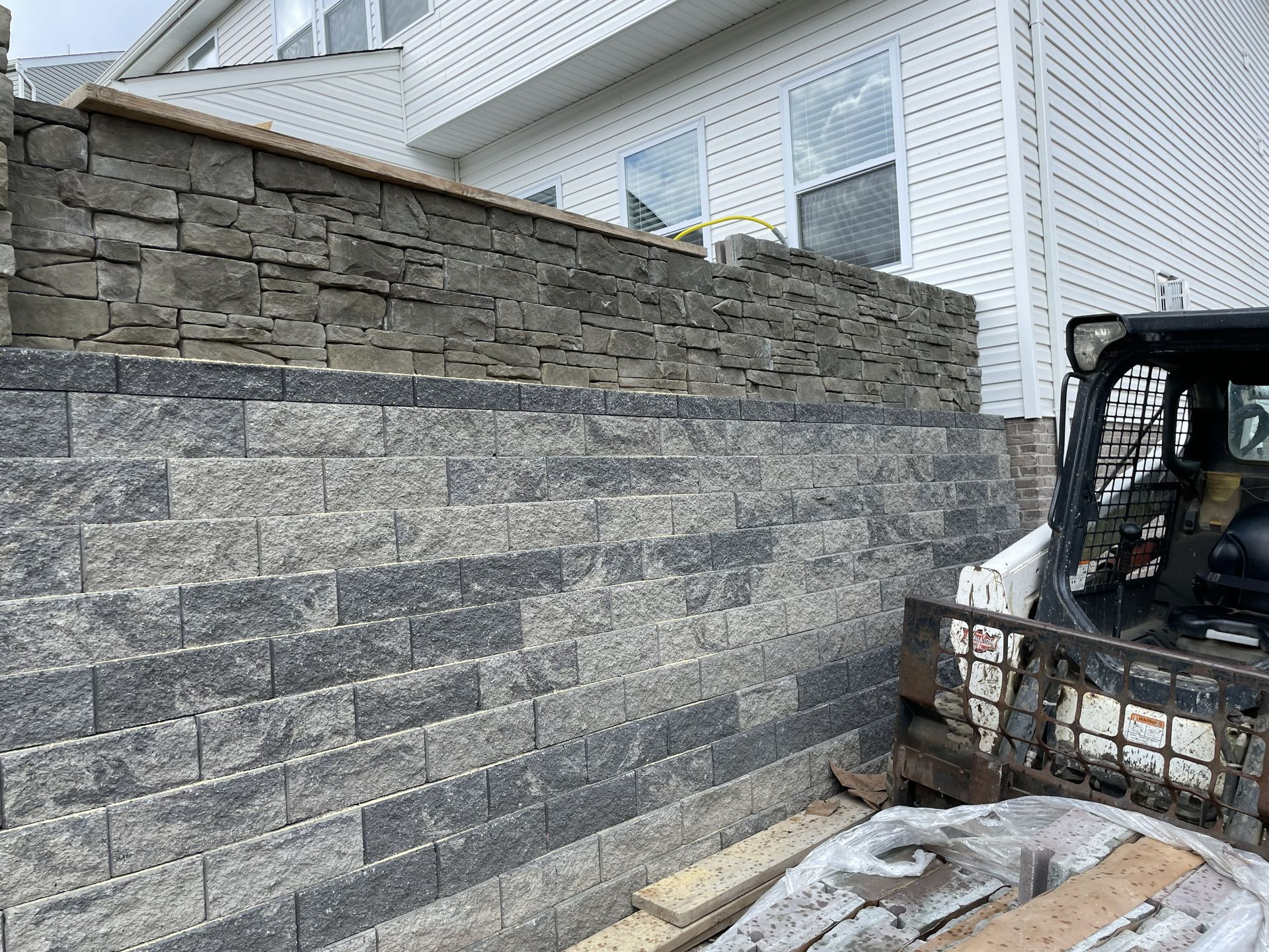 Gray stone retaining wall beside a white house, with a skid steer and snow on the ground