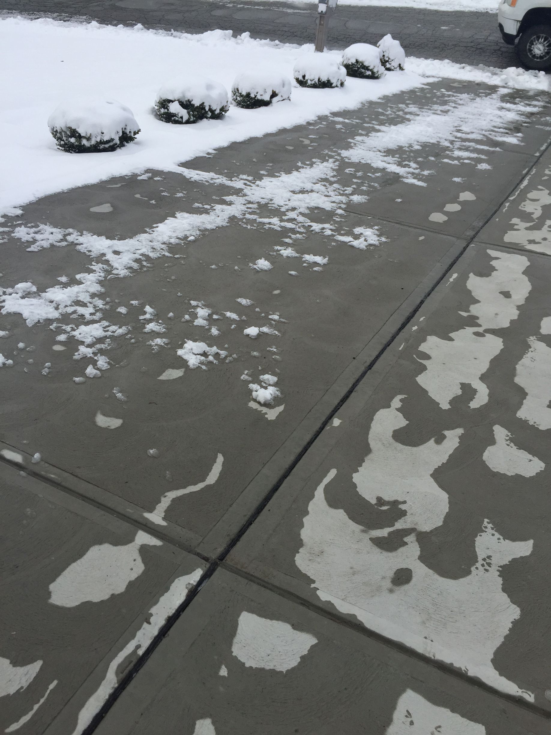 Snow-dusted concrete sidewalk and curb with patchy slush near a parking lot