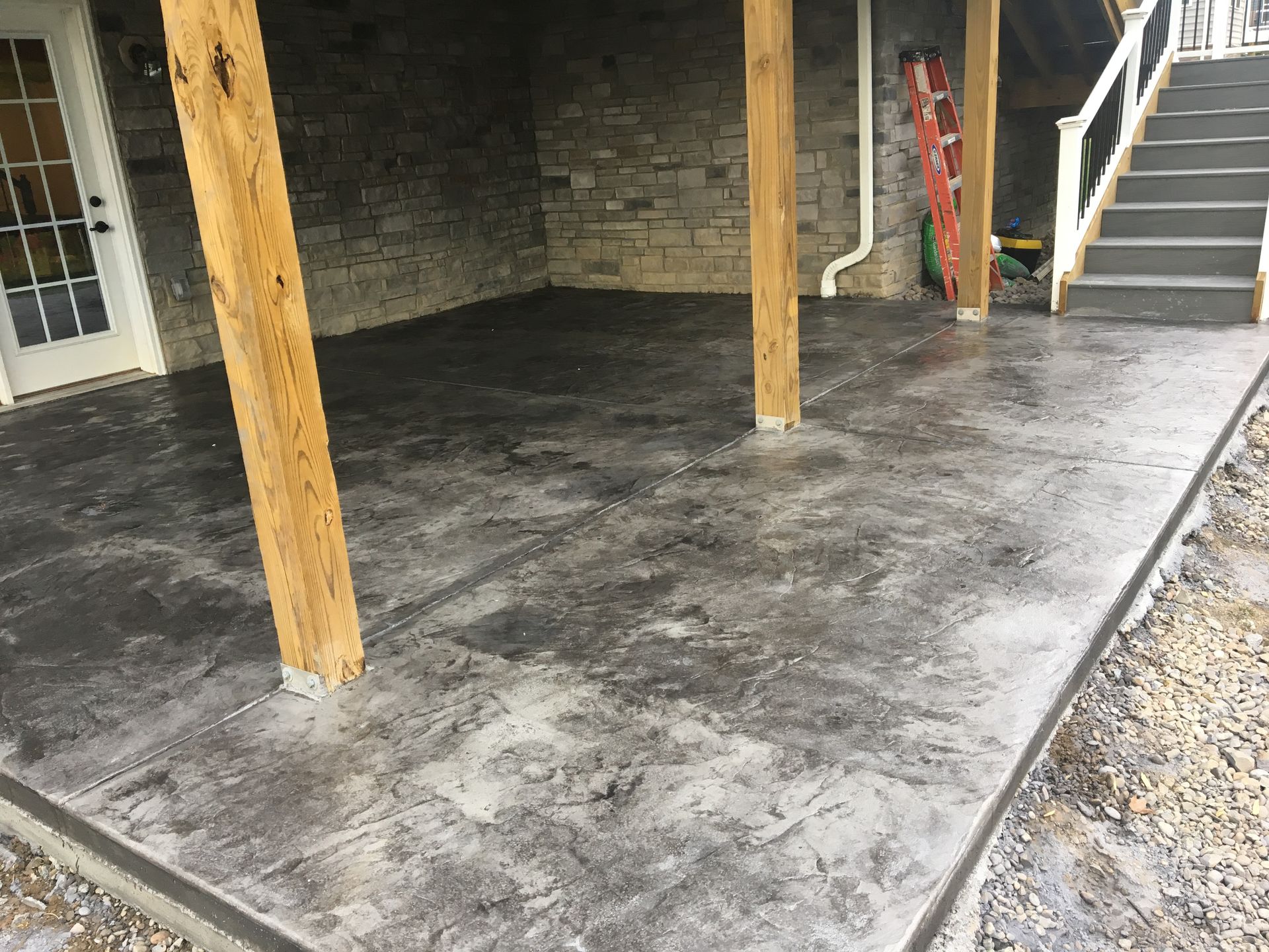Covered concrete patio with wooden posts, stone wall, and stairs leading up on the right.