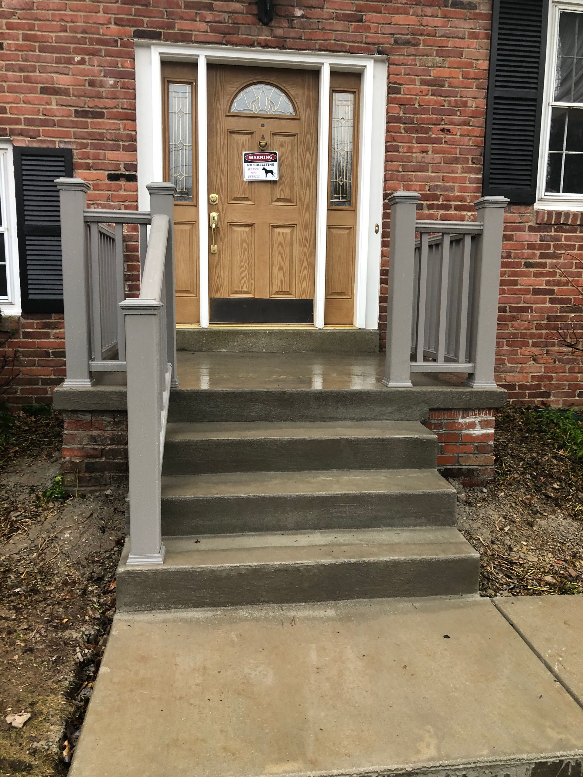 Brick house entrance with three concrete steps, gray railings, and a wooden door with a posted notice.