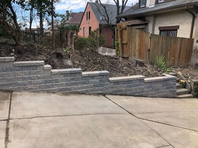 Stone retaining wall along a sloped yard beside a driveway, with houses and trees in the background