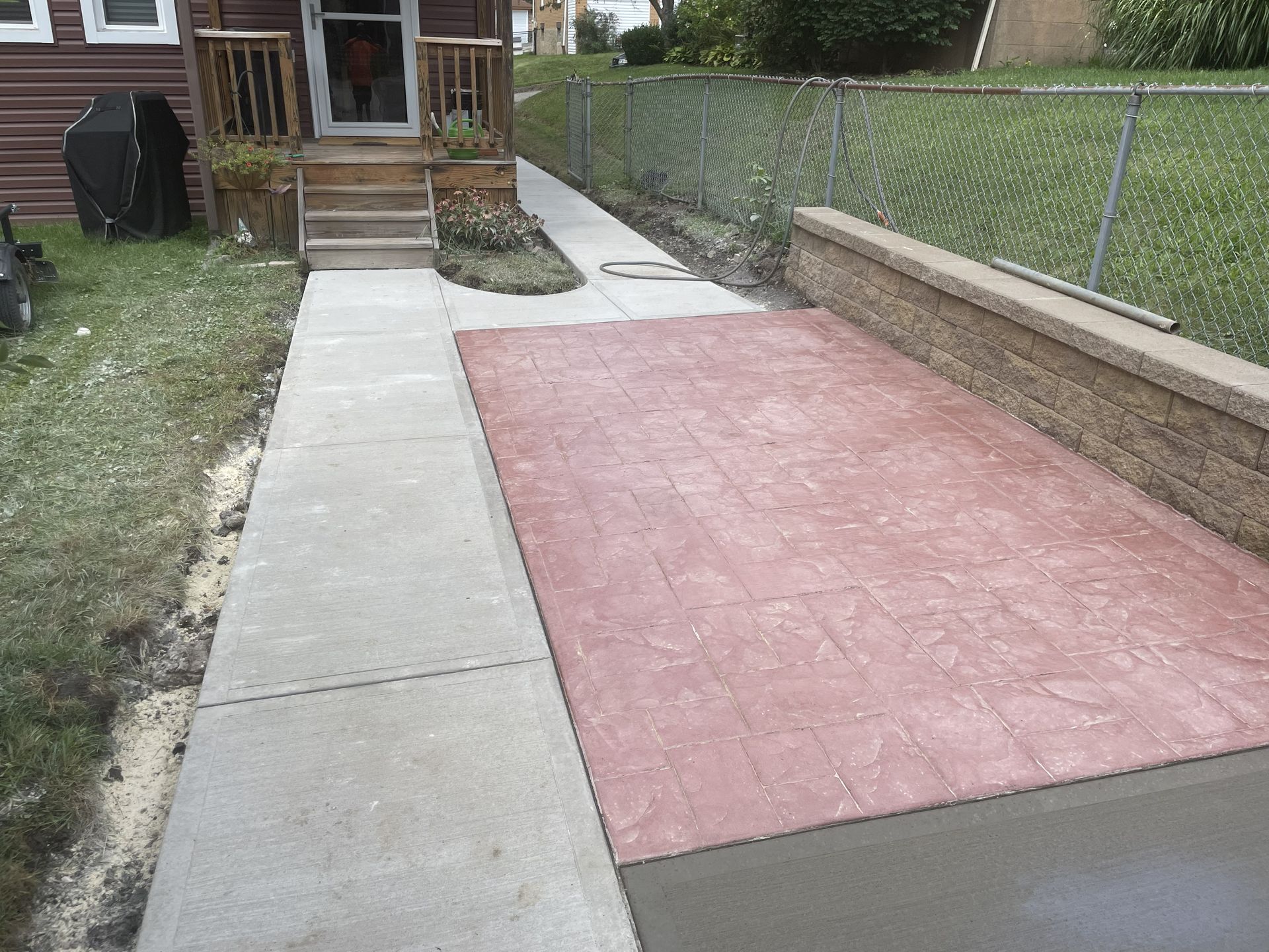 Concrete walkway beside a red stamped patio leading to a house entry, with a chain-link fence on the right.