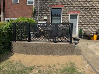 Small raised patio with black lattice fence and gate in front of a brick house.