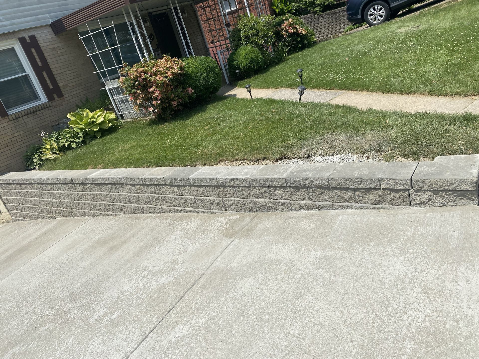 Sloped concrete driveway beside a grassy lawn and brick house with shrubs and parked car