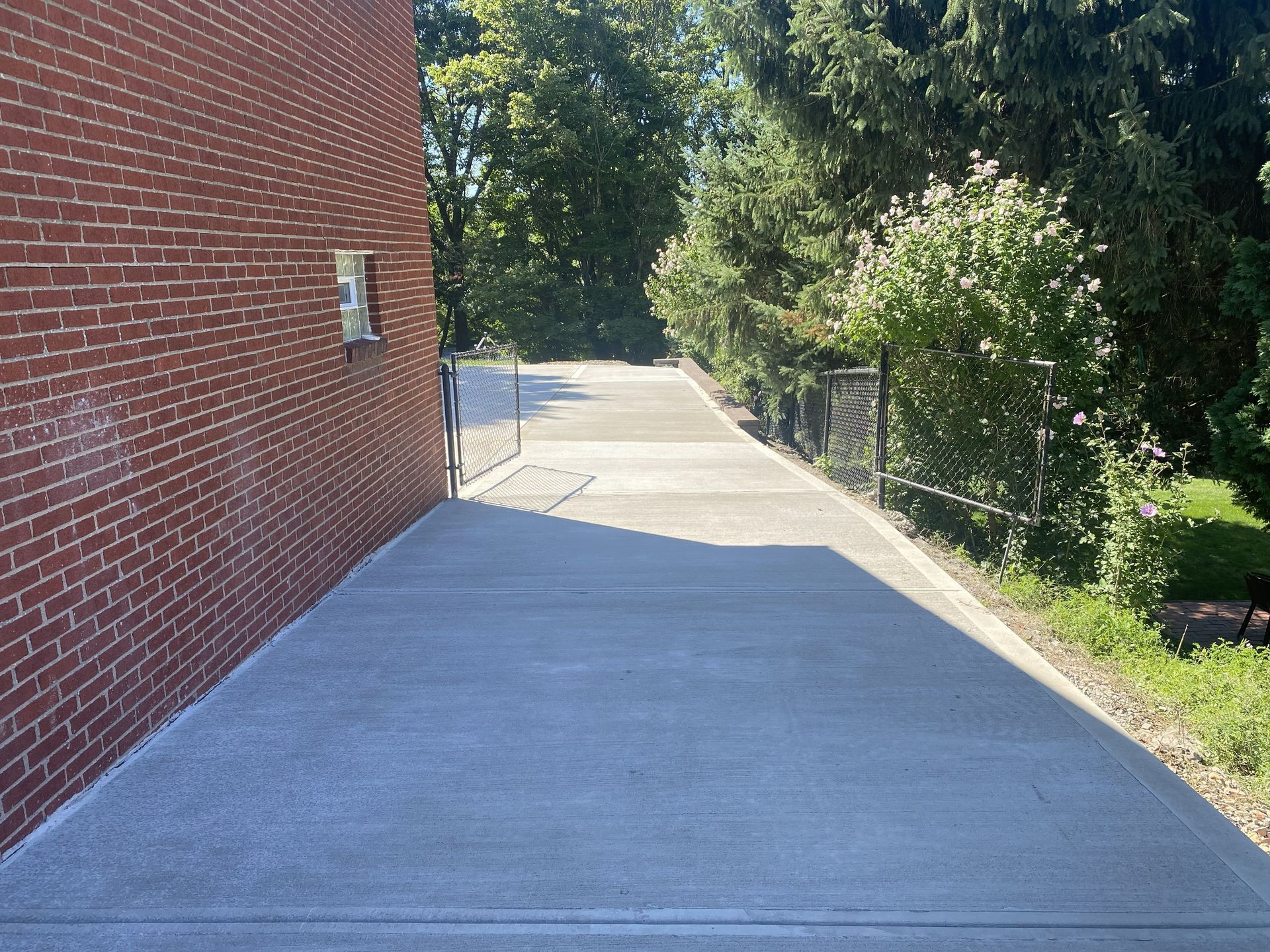 Concrete sidewalk beside a brick building, lined with trees and shrubs on a sunny day