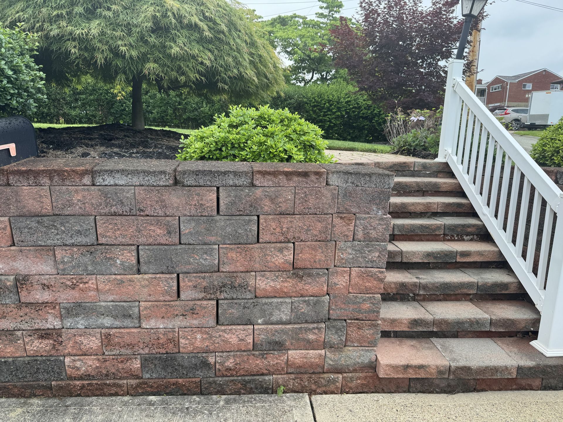 Brick retaining wall beside outdoor stairs with white railing and greenery behind.