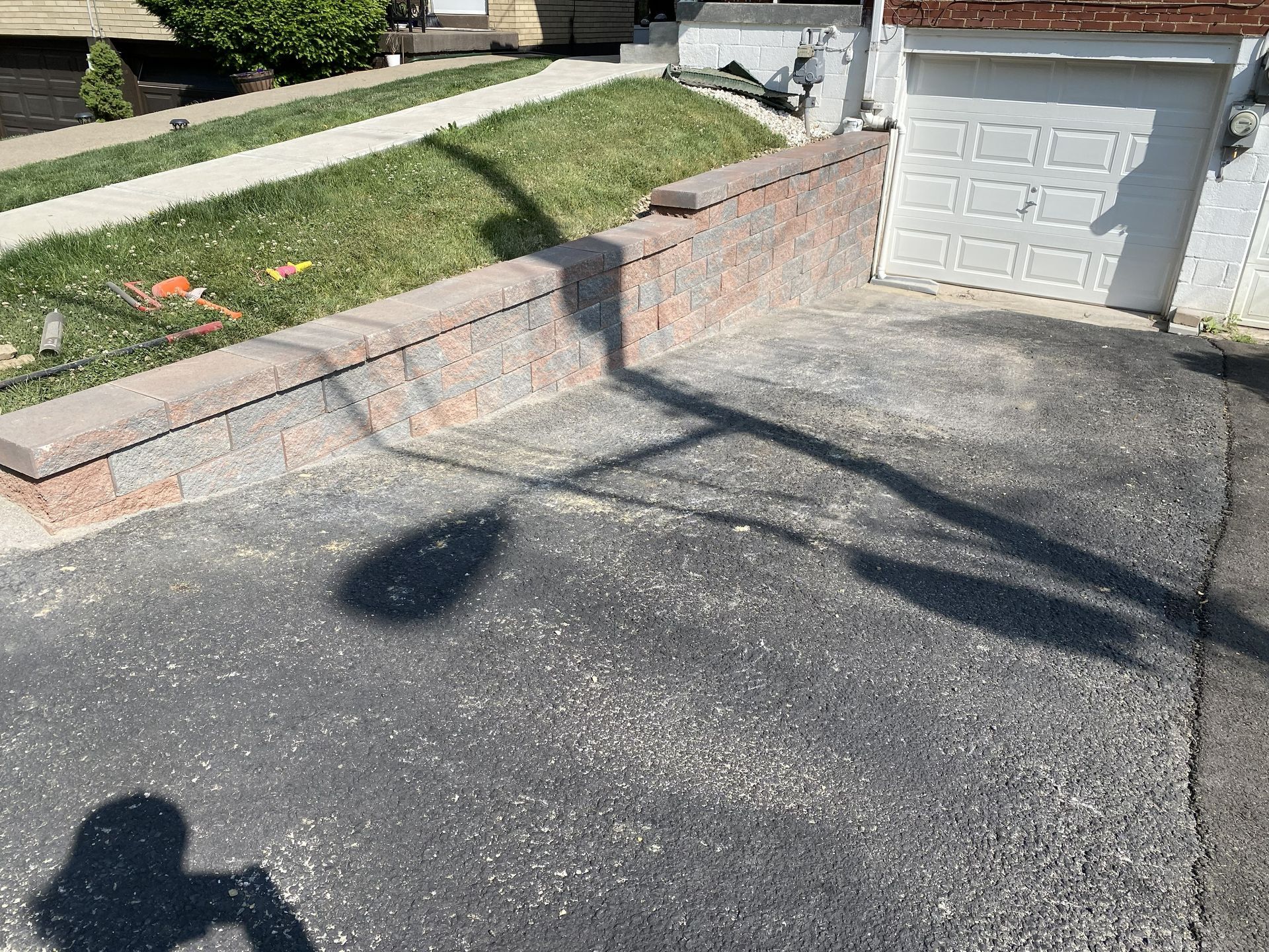 Driveway beside a sloped lawn and brick retaining wall, with a garage and tree shadows in sunlight.