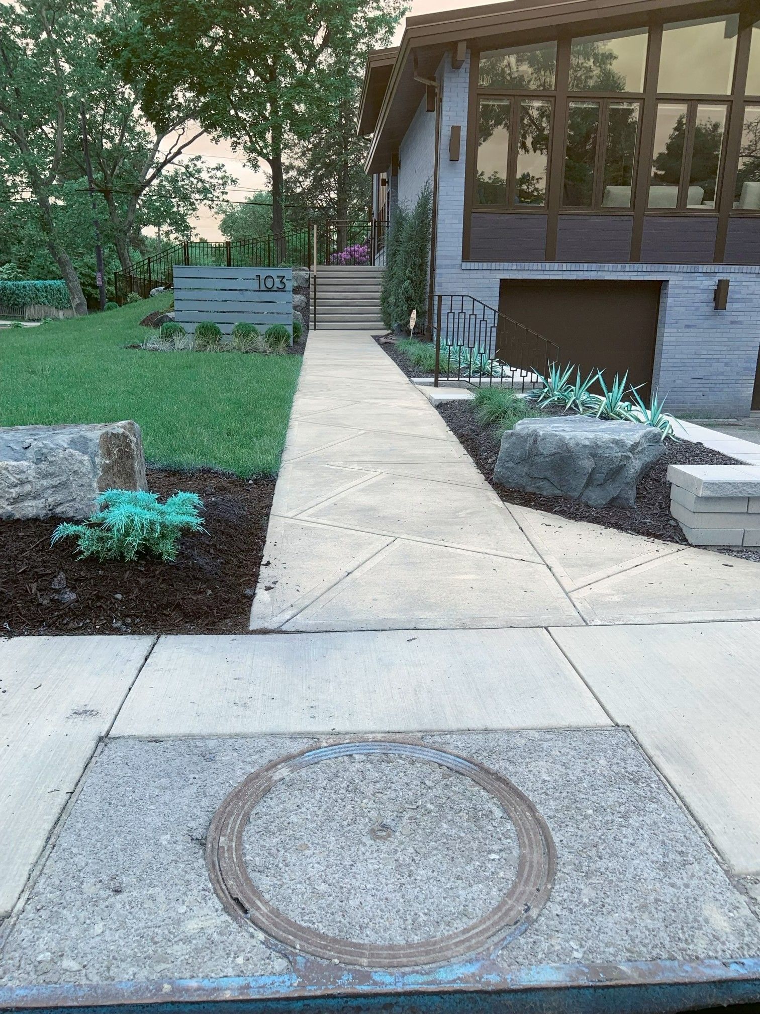 Concrete walkway leading to a house entrance with lawn, shrubs, and a raised planter bed