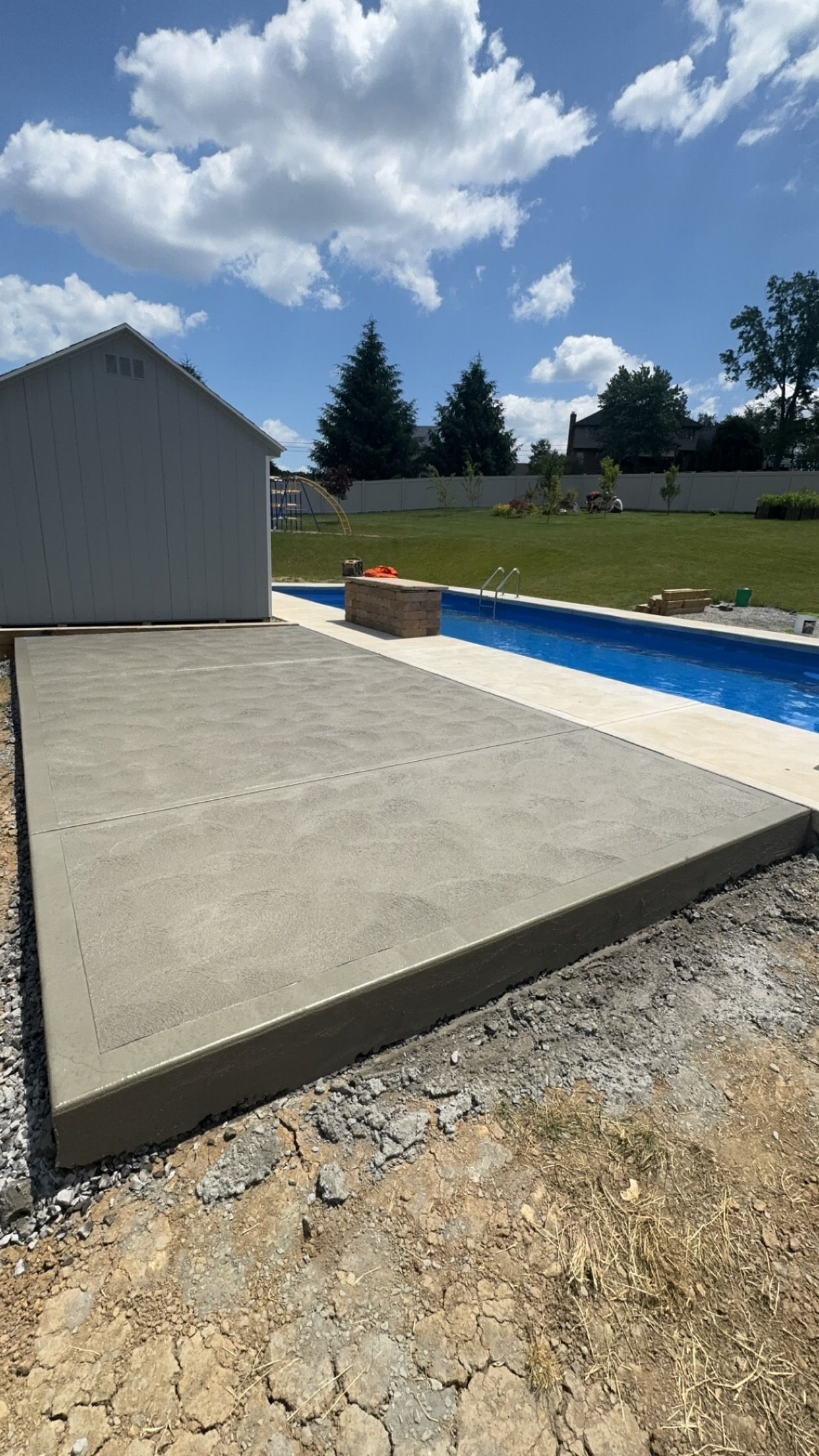 Concrete pool deck beside empty blue-lined pool, with gravel foreground, shed, and cloudy sky.