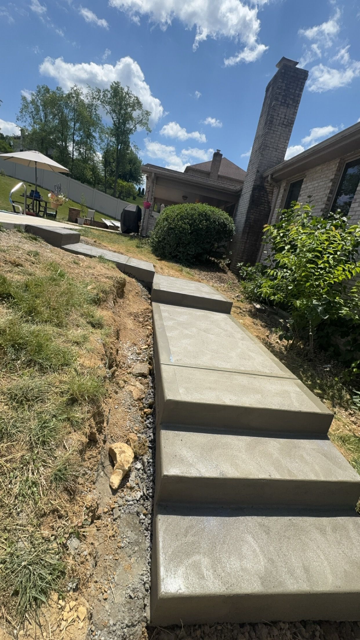 Concrete steps and sidewalk leading uphill past shrubs toward a house under a bright blue sky.