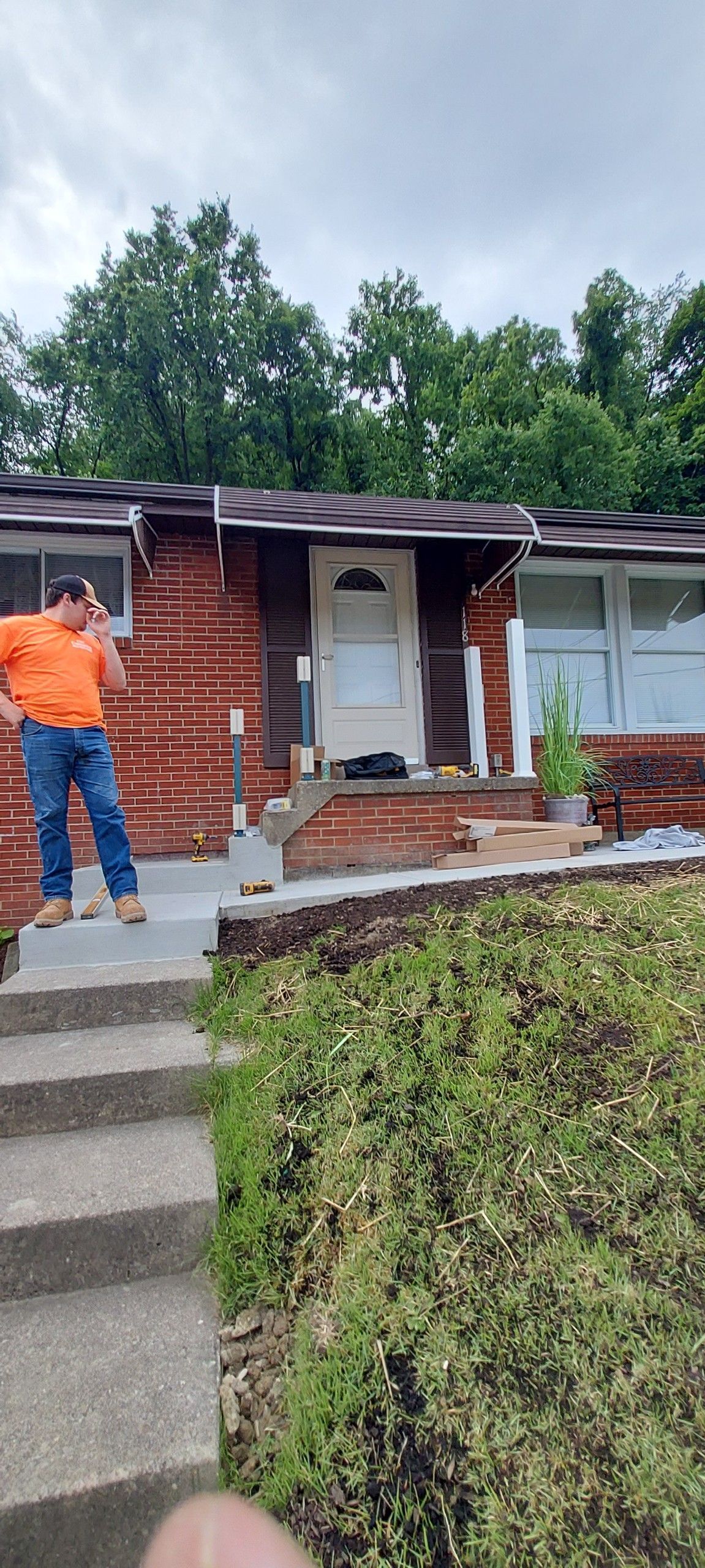 Person in orange shirt standing on front steps of a brick house with a white door and trimmed hedge.