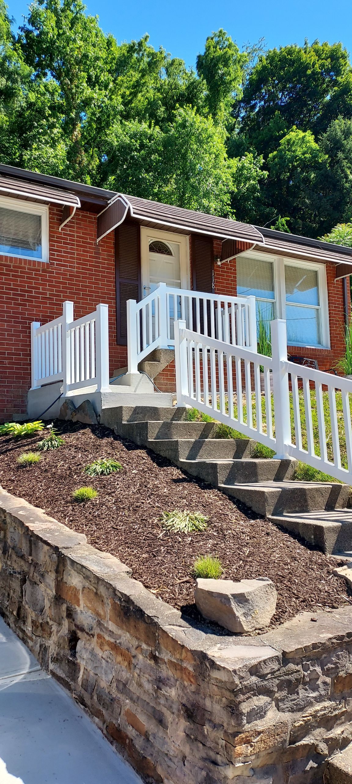 Red brick house with white railings and steps on a sloped stone retaining wall, surrounded by trees