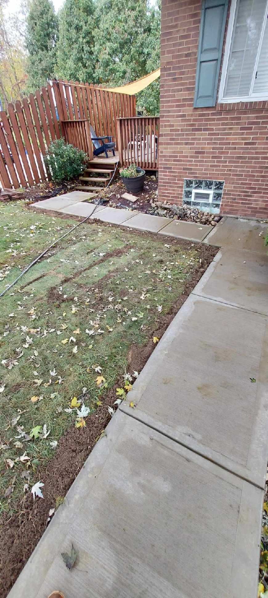 Small backyard patio beside a brick house, with a concrete walkway, wooden fence, and a potted chair area.
