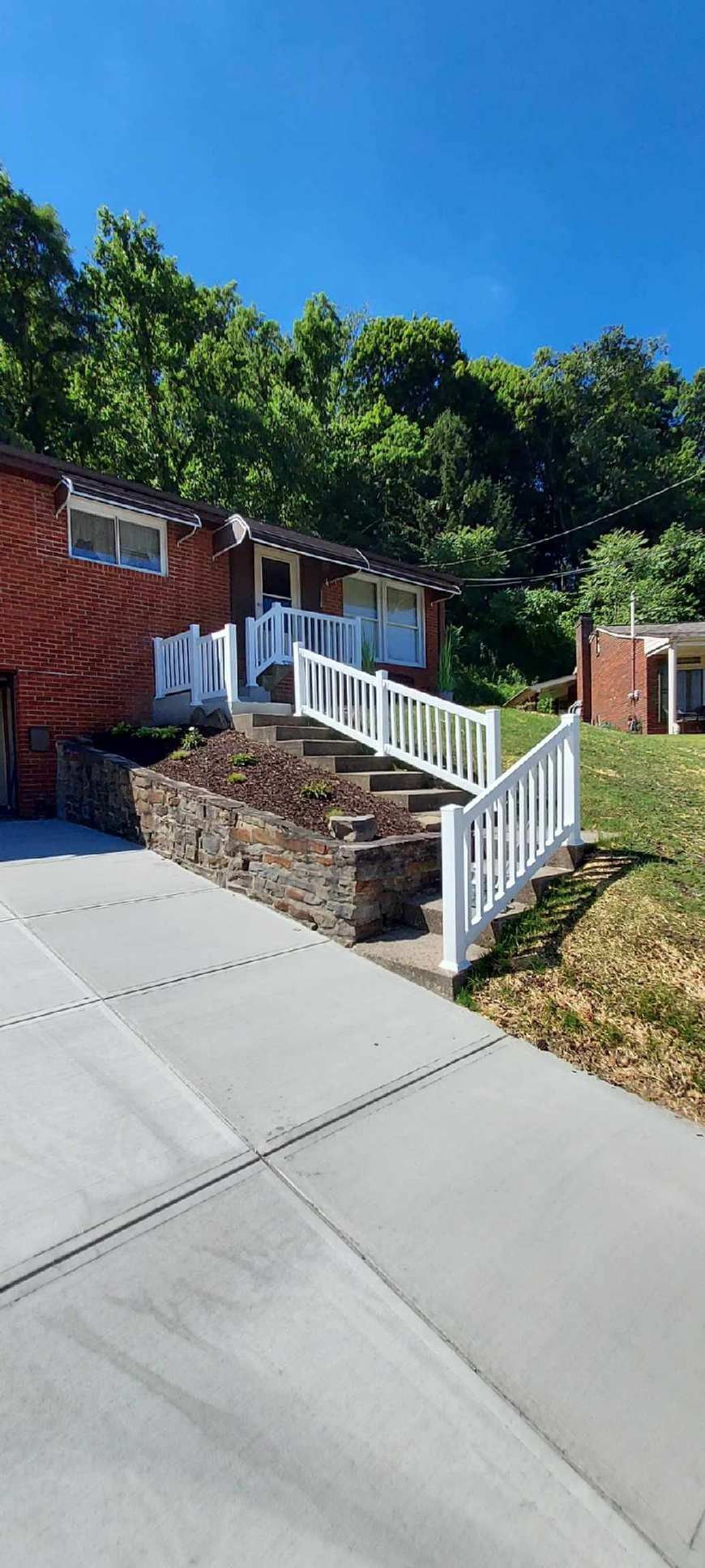 Brick house entrance with concrete steps and white railings beside a sloped driveway and trees