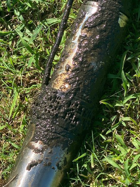 A corroded metal pipe covered in dark, oily residue lies on green grass.