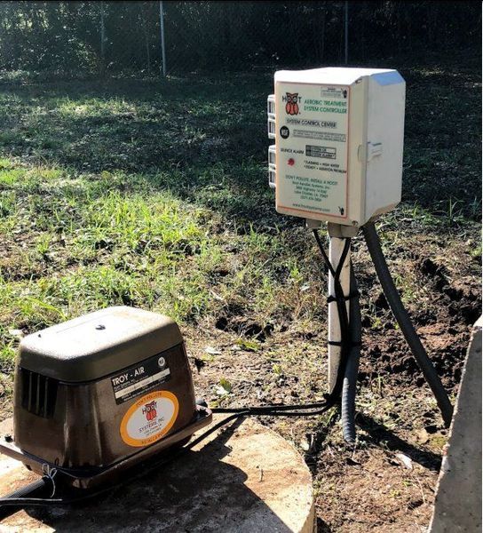 A brown pump and a white control box on a tripod stand outdoors near grass and a fence.