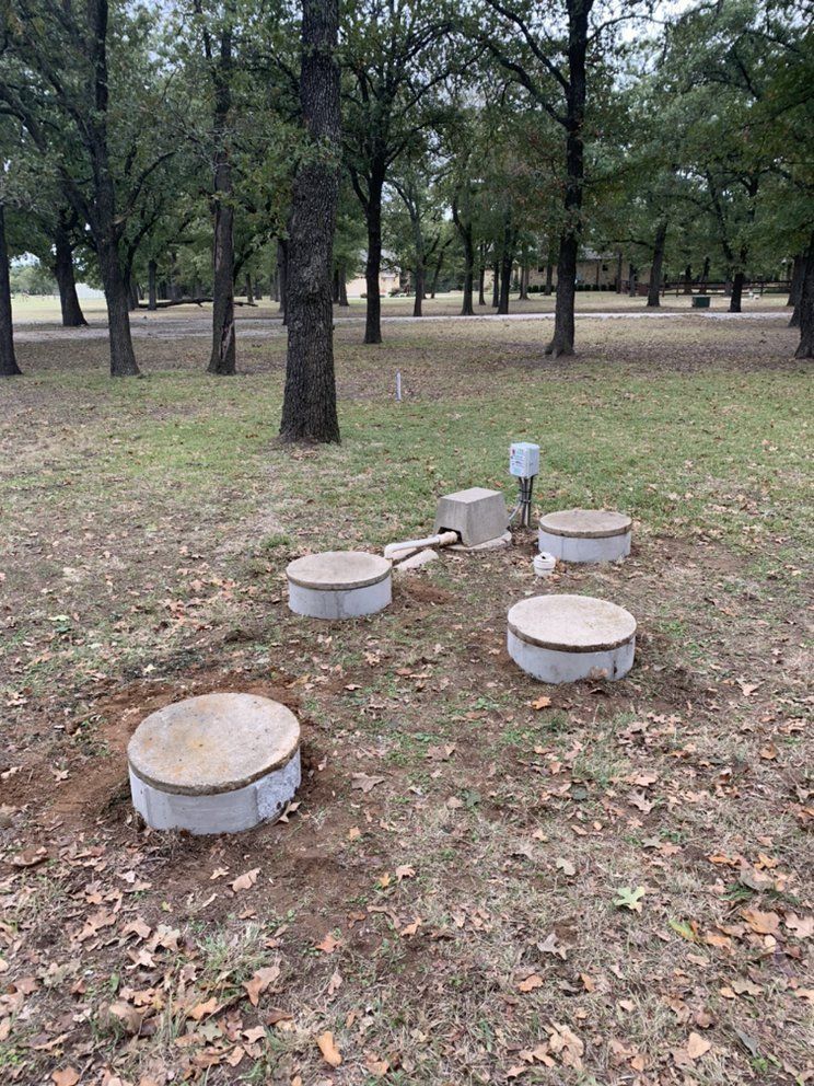 Septic system components in a grassy yard, including concrete lids and a control box, with trees in the background.