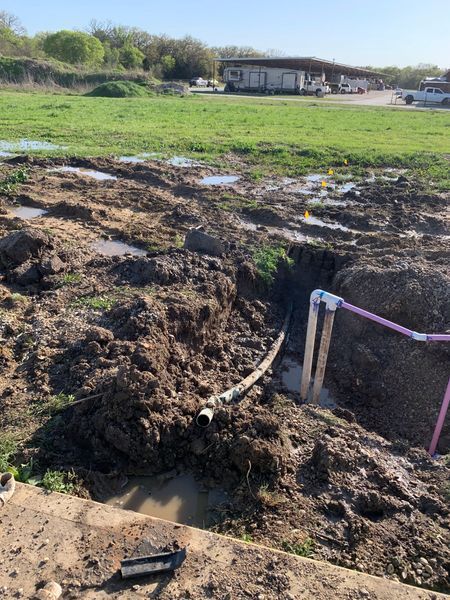 Muddy ground with exposed pipes, a ditch, and a building in the background. Pink pipes connect to a vertical pipe.