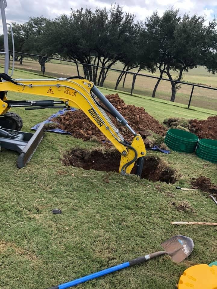 A yellow excavator digs a hole in green grass. Brown dirt piles surround the hole. Green containers and a shovel are nearby.