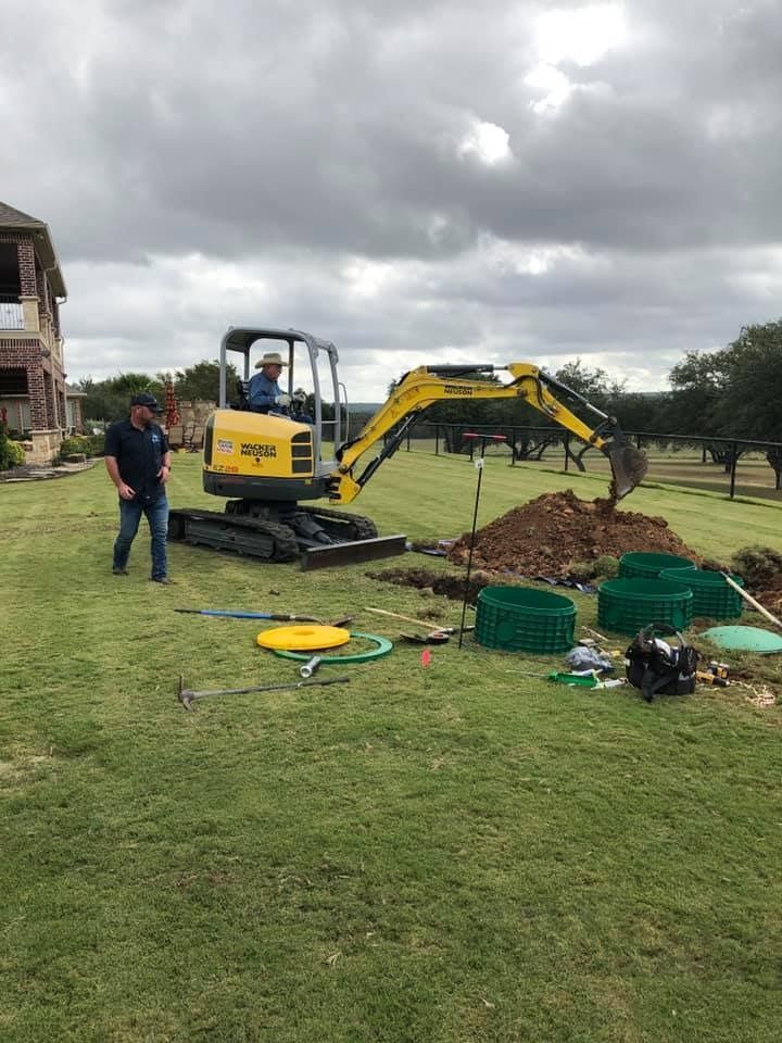 A small excavator digs in a grassy yard while a man stands nearby; several septic system components are visible.