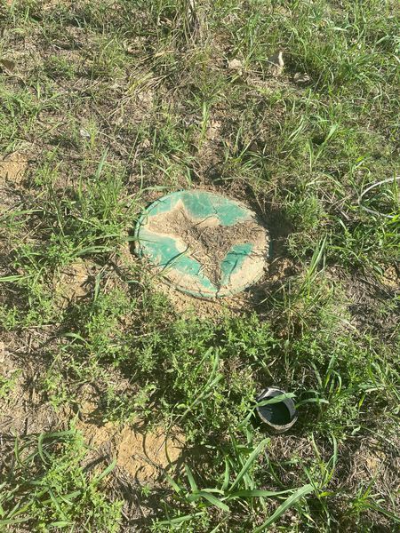 Green and brown circular cover in dirt and grass. Black object to the right.