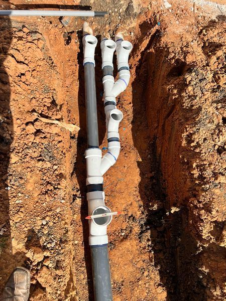 Plumbing pipes in a dirt trench, showing multiple Y-shaped connectors with gray and white PVC pipes.
