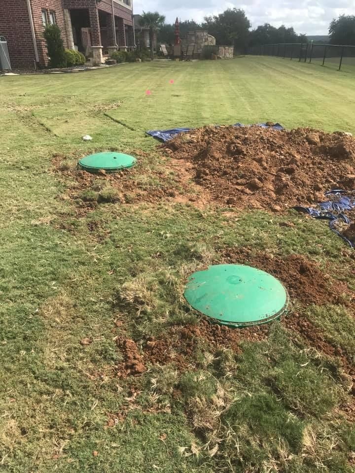 Two green septic tank covers in a grassy yard, with a dirt pile and a house in the background.
