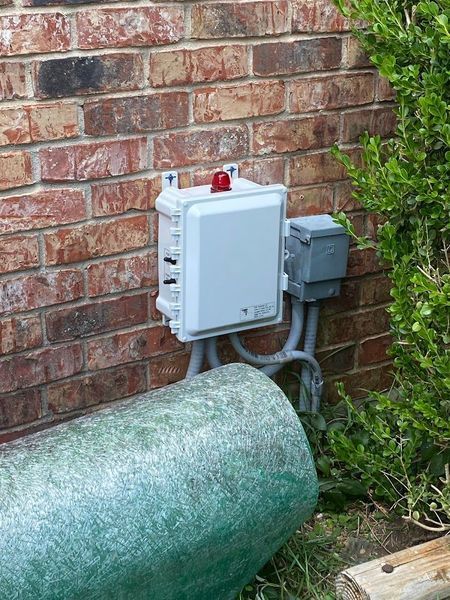 A gray utility box mounted on a brick wall with conduits and a red light on top.