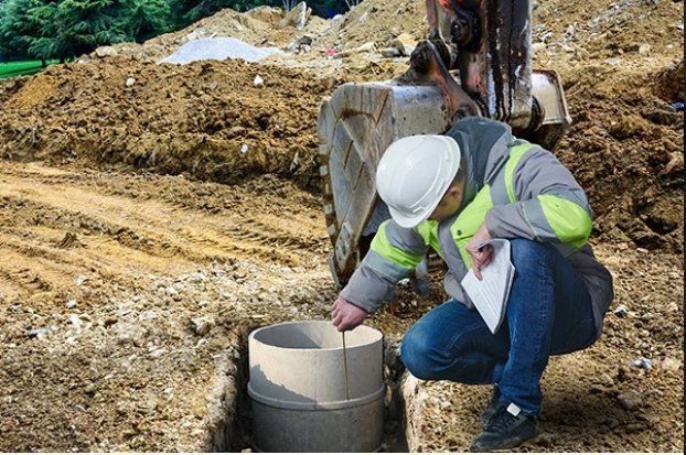 Construction worker inspecting a cylindrical concrete structure in a dirt excavation, wearing a hard hat and safety vest.