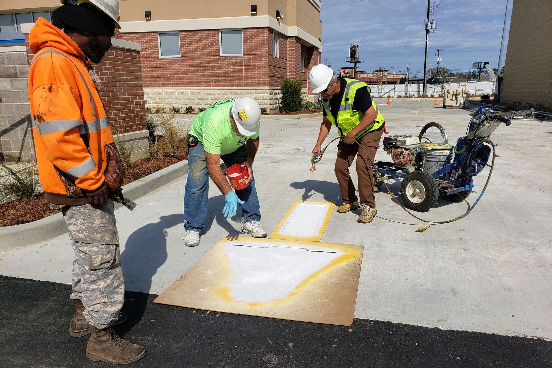 Workers painting curb markings on a concrete lot beside a brick building