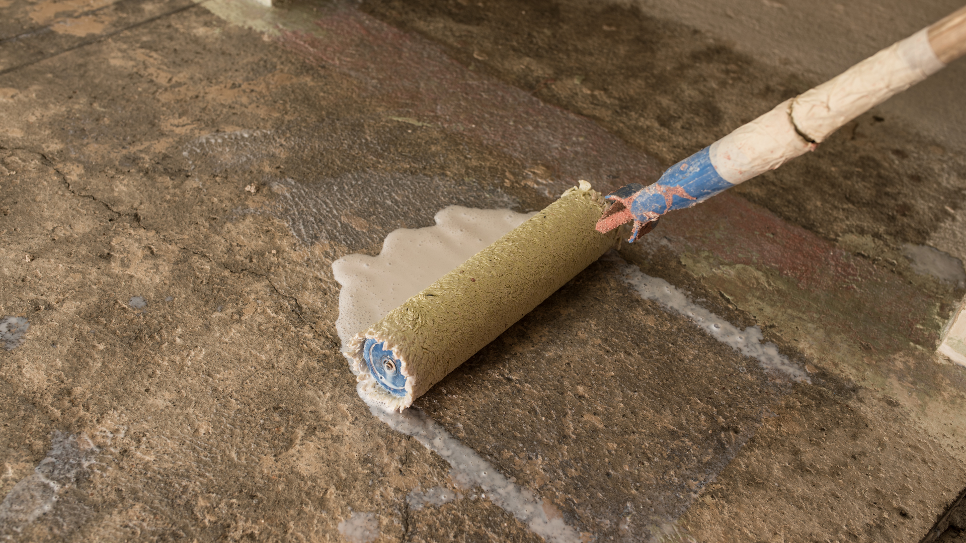 Paint roller applying white paint on a rough concrete floor near a wall line