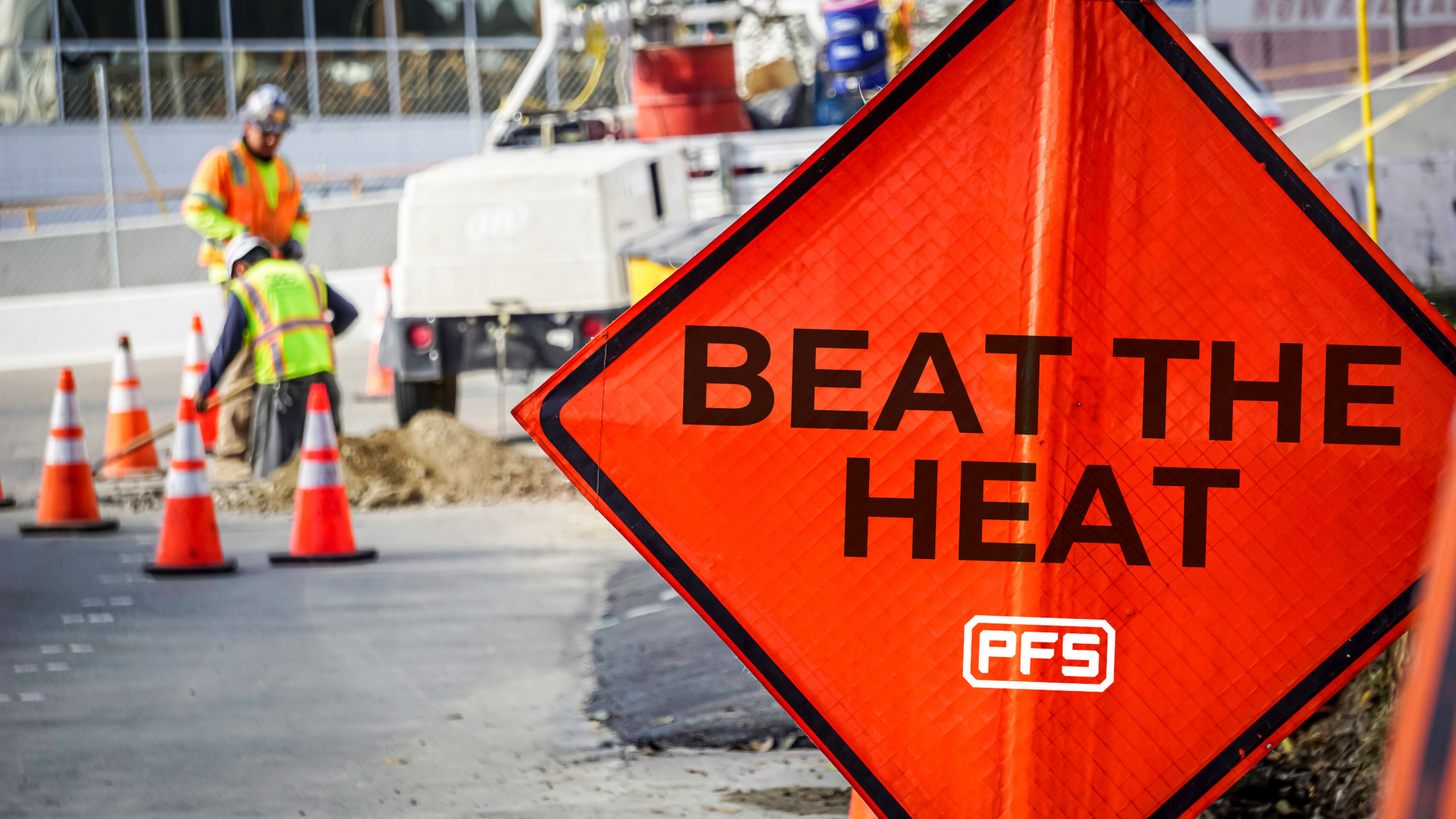 Orange roadwork sign reading “BEAT THE HEAT” beside traffic cones and workers on a street construction site