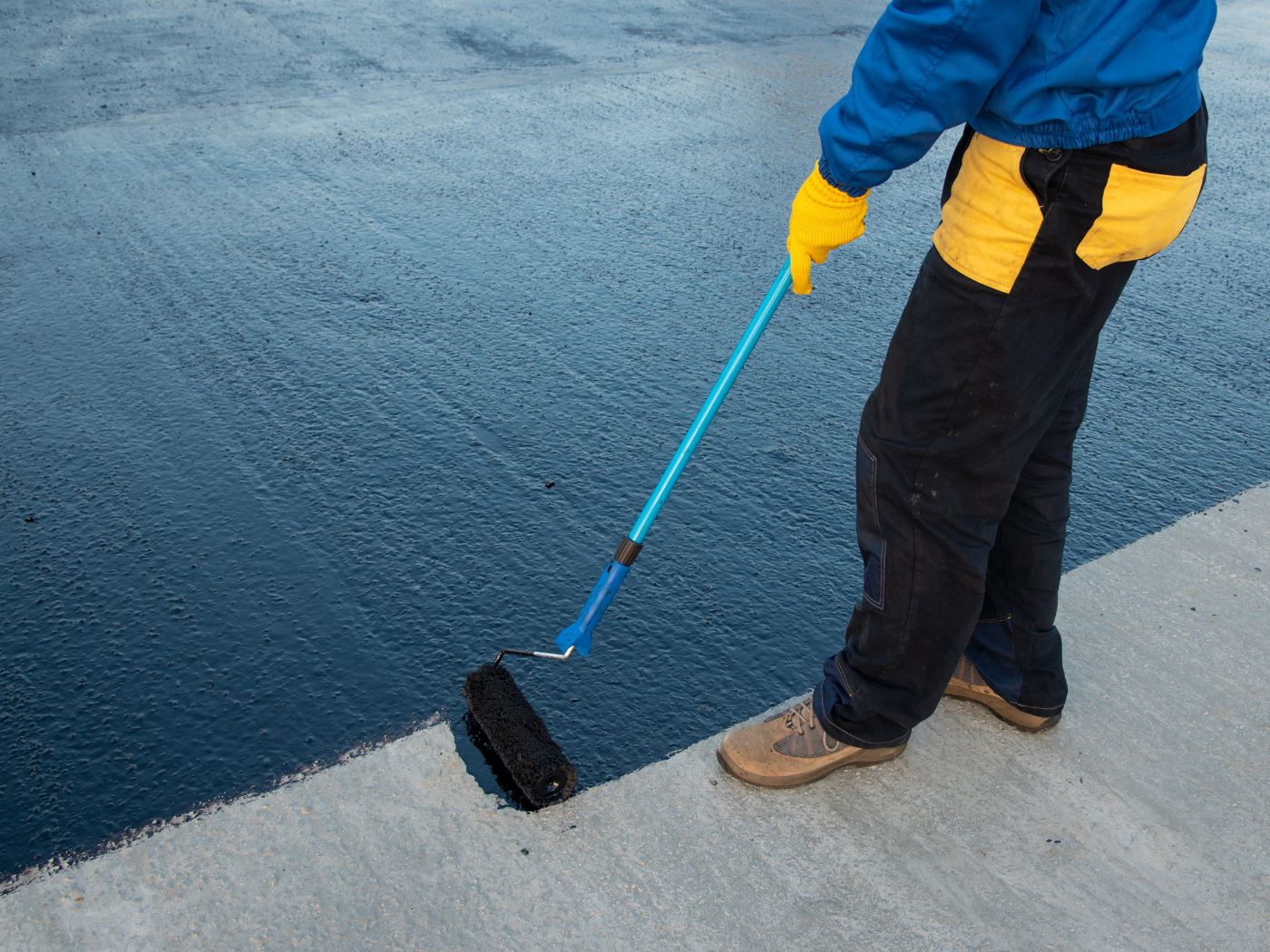 Person rolling black paint on pavement with a blue-handled roller, wearing blue jacket and yellow gloves.
